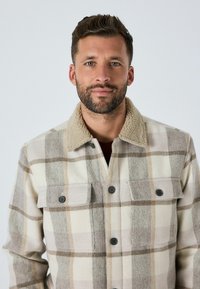 Flanelljacke mit einem karogenen Muster in Beige und Creme, ausgestattet mit einem weichen beige Sherpa-Kragen, zwei Brusttaschen und schwarzen Knöpfen.