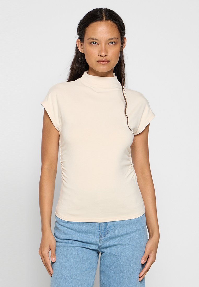Cremfarbener Mock-Neck-Top mit kurzärmeligen, gerafften Details an den Seiten, aus weichem, dehnbarem Stoff. Kombiniert mit hellblauen Jeans.