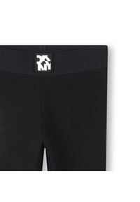 Leggings noirs avec une large ceinture élastique ornée d'un écusson logo. Le tissu semble lisse et extensible, conçu pour le confort et la flexibilité.