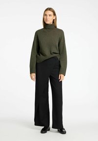 Khakifarbener Rollkragenpullover kombiniert mit schwarzen, weit geschnittenen Hosen mit asymmetrischem Design und dekorativen Knopfdetails. Strukturierter Strickstoff.