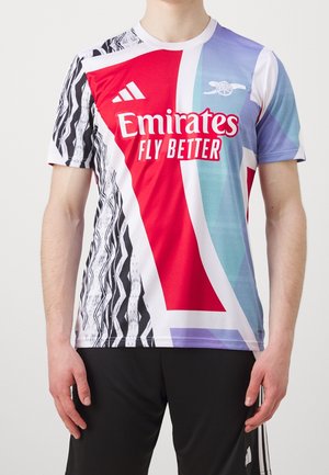 Person iført en farverig Arsenal fodboldtrøje med Emirates Fly Better-logo og Adidas-branding, kombineret med sorte Adidas-bukser.
