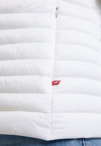 Biała pikowana kurtka od przodu z częściowo widocznym zamkiem błyskawicznym i małą czerwoną metką z tkaniny blisko dolnego brzegu, noszona na niebieskich dżinsach.