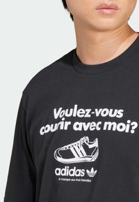 Fekete pamut póló, fehér felirattal: "Voulez-vous courir avec moi?", valamint egy Adidas cipő grafikájával. A póló kerek nyakkivágással és puha tapintással rendelkezik.