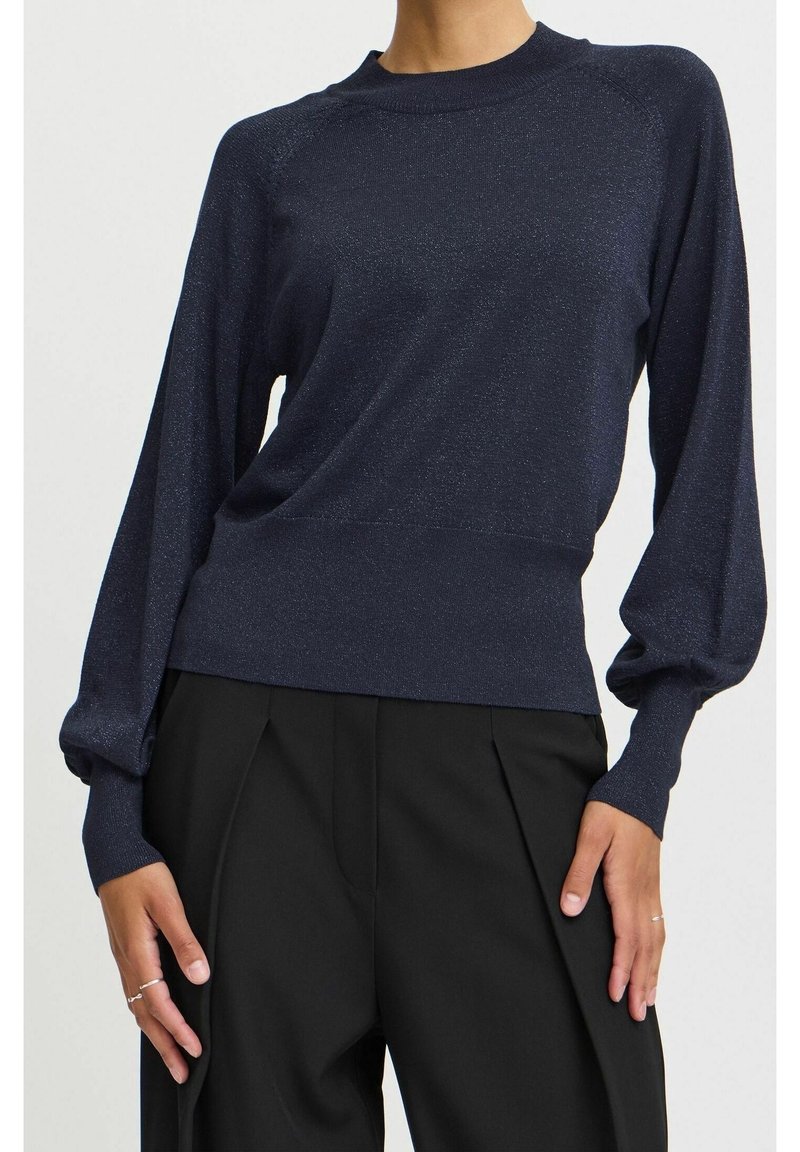 Modèle portant un haut à manches longues bleu marine foncé avec des manches bouffantes et des poignets ajustés, associé à un pantalon noir plissé taille haute.