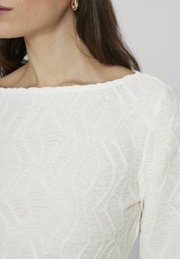 Blusa bianca con texture, caratterizzata da un motivo ondulato, scollatura off-shoulder e dettagli di leggero raccolto lungo i bordi.