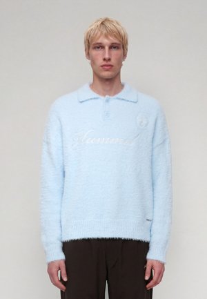 Homme blond portant un pull duveteux bleu clair avec un col et une broderie, debout devant un fond gris clair uni.