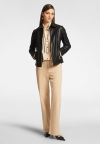 Giacca di pelle nera con zip frontale, blusa beige con decorazioni verticali nere, pantaloni beige chiaro e décolleté neri a punta.