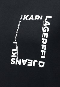 Μαύρο ύφασμα με λευκό κείμενο διατεταγμένο σε τετράγωνο: «KARL LAGERFELD JEANS» επαναλαμβανόμενο γύρω από τις άκρες.