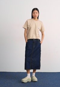 T-shirt beige à poche, jupe midi en jean foncé, sabots vert clair, chaussettes blanches. Design simple, coupe décontractée, coutures visibles sur la jupe.