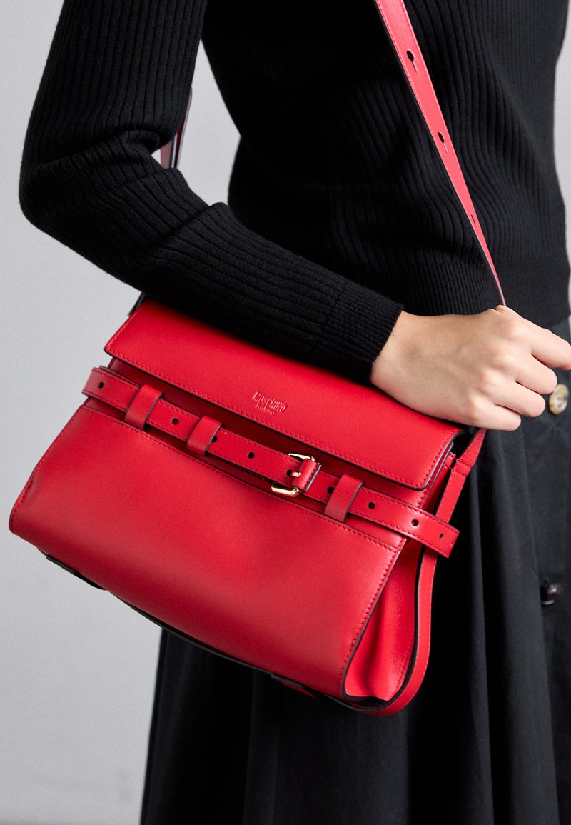 Woman wearing black long-sleeve top and skirt holds a structured red leather crossbody bag with buckle detail and adjustable strap.