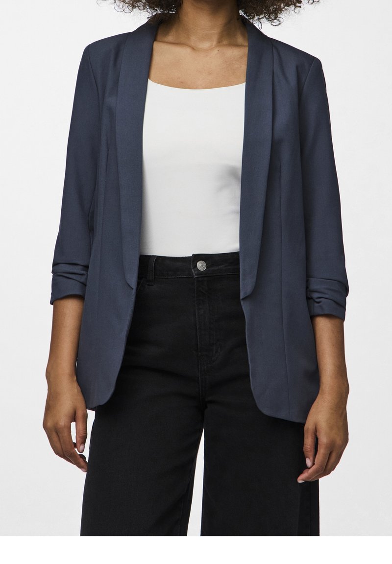 Blazer bleu foncé à texture lisse, ouvert sur le devant avec les manches retroussées. Porté sur un haut blanc et associé à un pantalon taille haute noir.
