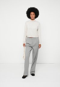 Woman standing against white background wearing a light beige turtleneck, gray wide-leg pants, black shoes, and holding a beige handbag.
