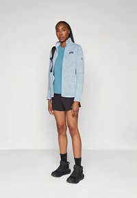 Hele sinine fliisjakk kõrge kraega, mida kannab türkiissinine särk ja mustad lühikesed püksid. Mustad matkajalatsid ja seljakott täiendavad rõivastust.