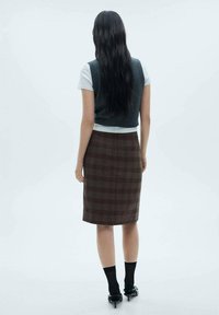 Vest gri tricotat peste un tricou alb, asociat cu o fustă creion cu carouri maro, șosete negre și pantofi cu toc ascuțit negri. Fond minimalist.