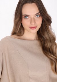 Lys beige sweater med en blød ribbet tekstur, bred bådhals og diagonalt strikket mønsterdetalje på den ene side. Glat stoffinish.