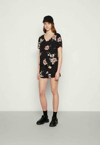 Top curto de manga curta com padrão floral preto e decote em V, combinado com calções florais iguais. Modelo a usar uma boné preto e sapatos escuros.