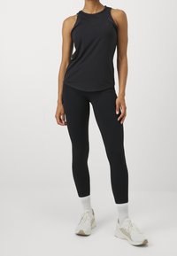Černý bezrukávový sportovní top s zaobleným lemem a výřezy na ramena, kombinovaný s černými legínami a bílými teniskami s béžovými akcenty.