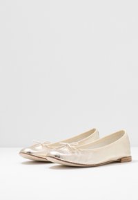 Bailarinas metálicas de color dorado con parte superior de cuero suave, puntera redondeada y lazo decorativo en la parte frontal. Suela plana con tacón mínimo.