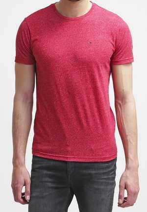 Man draagt een nauwsluitend rood T-shirt met korte mouwen en donkergrijze spijkerbroek, staand tegen een effen witte achtergrond.
