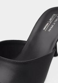 Μαύρο ανοιχτό παπούτσι τύπου mule με χαμηλό τακούνι, λείο δερμάτινο επάνω μέρος και ορατό κείμενο της μάρκας στη σόλα, σε λευκό φόντο.