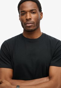 Zwarte katoenen t-shirt met korte mouwen en een ronde hals, aansluitend ontwerp met een zachte textuur. Armen gekruist in een neutrale houding.