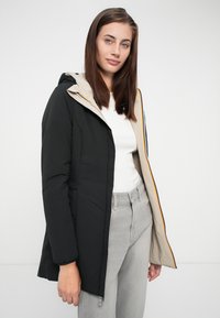 Zwarte gewatteerde jas met een lichtbeige voering en capuchon, voorzien van een rits aan de voorkant en zijzakken. Draagt over een witte top en grijze broek.