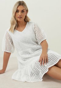 Vit klänning med en texturerad, öppen vävdesign, korta puffärmar och en volangkant. V-ringning. Modell sitter, med handen på knät.