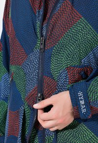 Ruka držící zip barevné bundy s modro-červenou-zelenou a světle modrou čárkovanou vzorovou tkaninou a textem "HYRA" na rukávovém štítku.