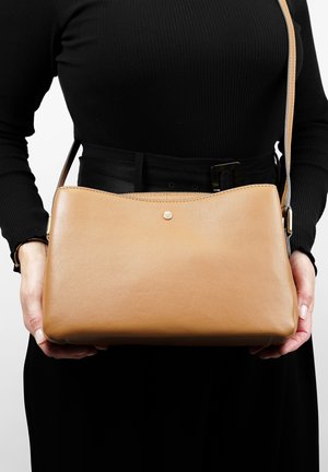 Tan leather crossbody bag with a seamless design, rounded edges, and a small gold accent on the front. Strap and texture visible.