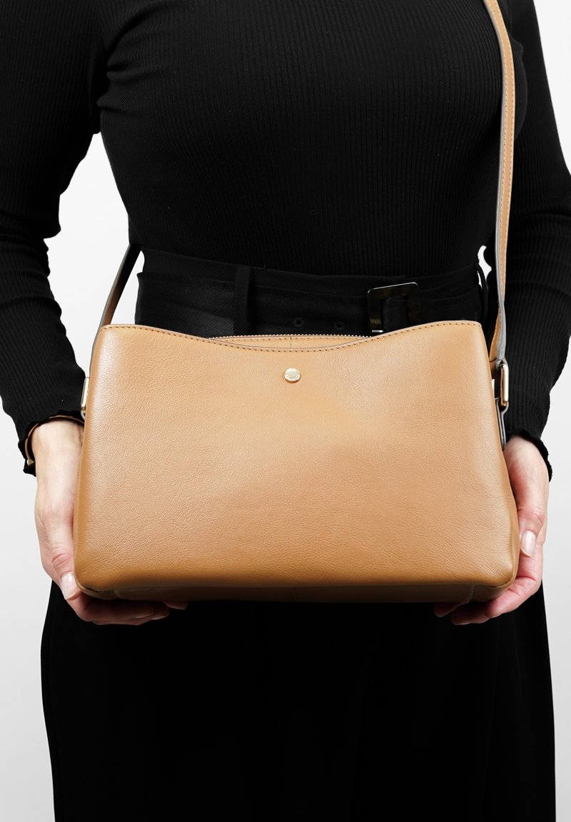 Tan leren schoudertas met een naadloos ontwerp, afgeronde randen en een kleine gouden accent aan de voorkant. Band en textuur zichtbaar.