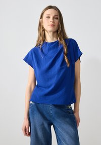 Maglietta blu con texture, maniche corte, scollatura rotonda e vestibilità ampia. Abbinata a jeans in denim blu chiaro.