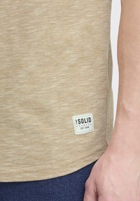 Tricou texturat bej cu margine rotunjită, având un patch cu logo alb și negru în partea stângă jos. Materialul pare moale și casual.