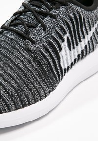 Czarno-białe skarpetowe buty sportowe z czarnymi sznurówkami i białą podeszwą, przedstawiające zbliżenie na wzorzystą tkaninę i szycie.