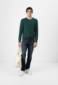 Pull en maille vert à manches longues, jeans bleus, baskets blanches, tenant un sac fourre-tout beige. Design simple, avec un détail de logo sur le pull.