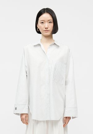 Femme portant une chemise blanche oversize à boutons avec fines rayures verticales, grande poche rayée et larges poignets, debout devant un fond uni.