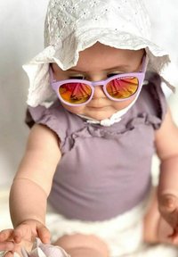 Lilatvärgade solglasögon med reflekterande orange linser, vit broderad solhatt och lätt lila ärmlös topp. En baby sträcker sig efter en blomma.