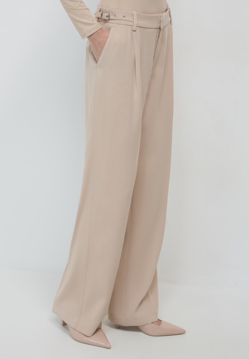 Pantalons larges beiges à taille haute, avec des plis et des passants de ceinture. Tissu lisse, comprenant des poches avant et un éclat subtil.