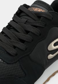 Chaussure de sport noire en matériau synthétique texturé, avec six œillets pour lacets et semelle blanche contrastante. Comprend un détail logo métallique.