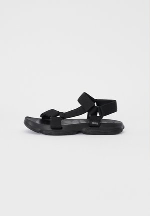 Sort sandal med gummisål, justerbare stofremme og tekstureret polstring. Minimalistisk design med et logo på hælen.