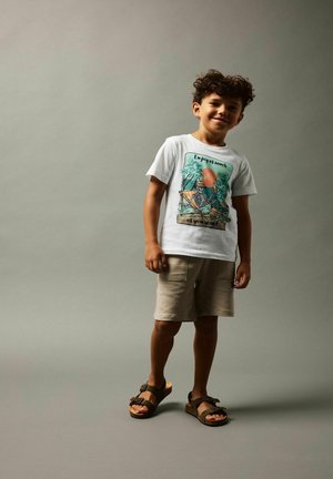 Camiseta blanca con estampado colorido que presenta un esqueleto y palmeras, combinada con pantalones cortos beige y sandalias marrones. Tejido de algodón suave.