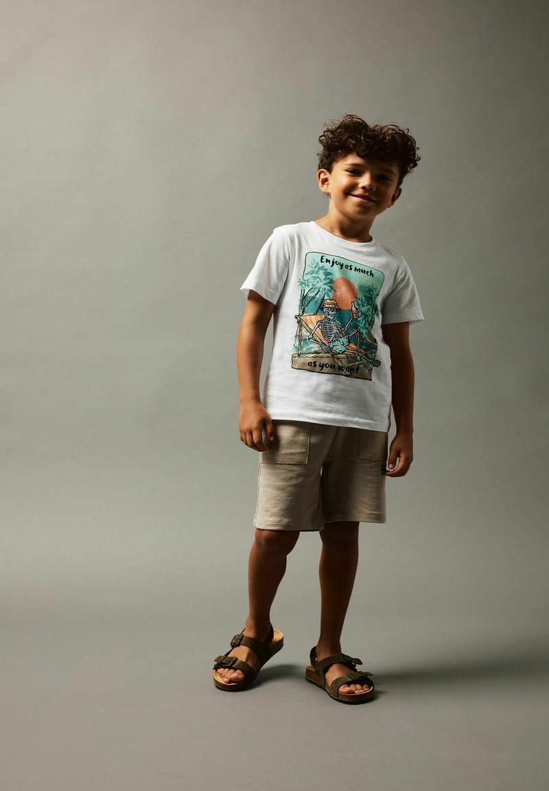 Camiseta blanca con estampado colorido que presenta un esqueleto y palmeras, combinada con pantalones cortos beige y sandalias marrones. Tejido de algodón suave.