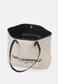 Canvas tote bag in lichtbeige met zwarte leren accenten, met een bovenste drukknoopsluiting en een binnenzak met rits. Branding zichtbaar aan de voorkant.