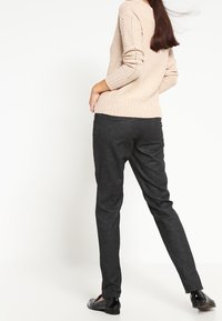 Frau in einem beigen Strickpullover, dunkelgrauen schmalen Hosen und schwarzen glänzenden Loafers, die mit dem Rücken zugewandt steht und eine Hand in der Tasche hat.