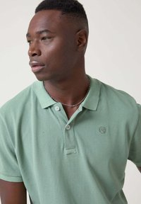 Polo de tela verde suave, con una tapeta de dos botones, mangas cortas y un sutil detalle de logo en el pecho.