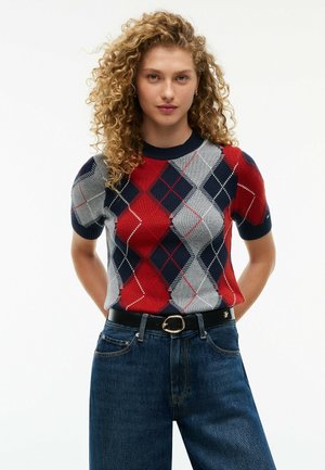 Lockige Frau trägt einen rot-grau-marineblauen Argyle-Kurzarmpullover, in blaue Jeans mit schwarzem Gürtel gesteckt.
