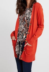 Oranje gebreide cardigan met twee vo pockets en houten knopen, gedragen over een gestreept top. Model draagt donkere jeans.
