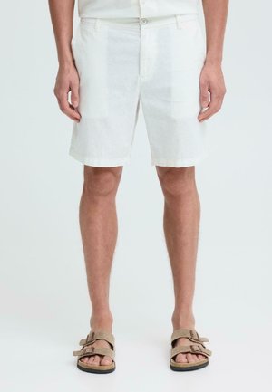 Mann iført hvite shorts og beige sandaler med to stropper, stående på en enkel lys bakgrunn.