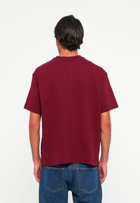 Hombre con cabello oscuro que lleva una camiseta de manga corta lisa de color granate y jeans azules, de espaldas contra un fondo blanco liso.