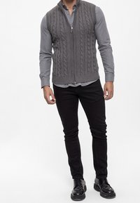 Grijze kabelgebreide mouwloze trui met rits, gedragen over een lichtgrijze overhemden, gecombineerd met zwarte slim-fit broek en zwarte schoenen.