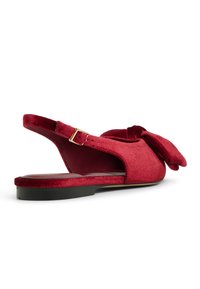 Rød fløjl slingback sko med flad sål, prydet med en stor sløjfe, justerbar spænde og åben bagside. Glat tekstur.